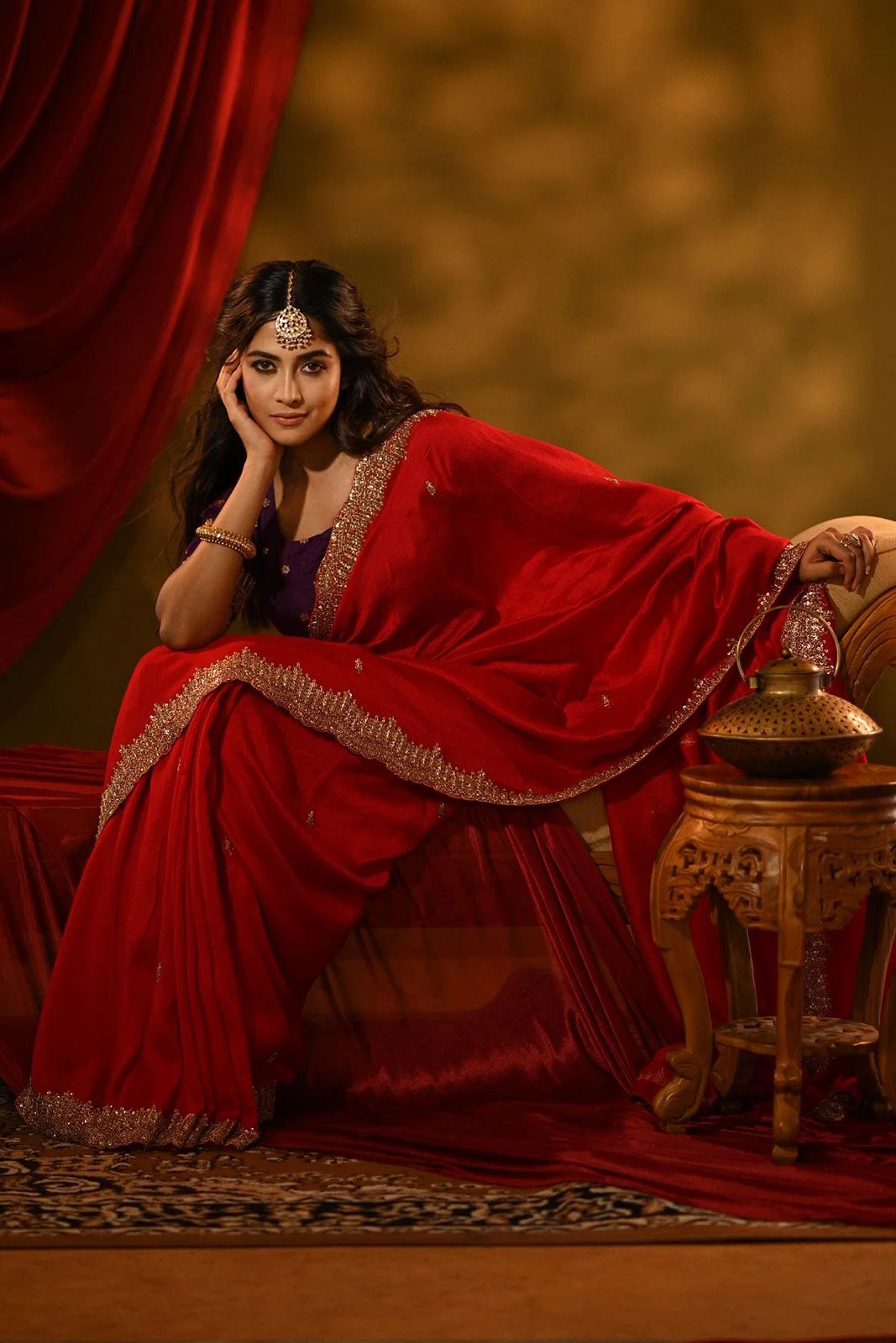 A woman in a red dress with a gold headpiece stands indoors near a red curtain. DesiColorClothing.com